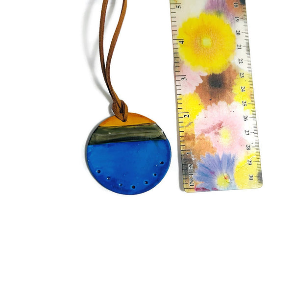 Round blue pendant with a brown cord next to a ruler with floral design on a white background