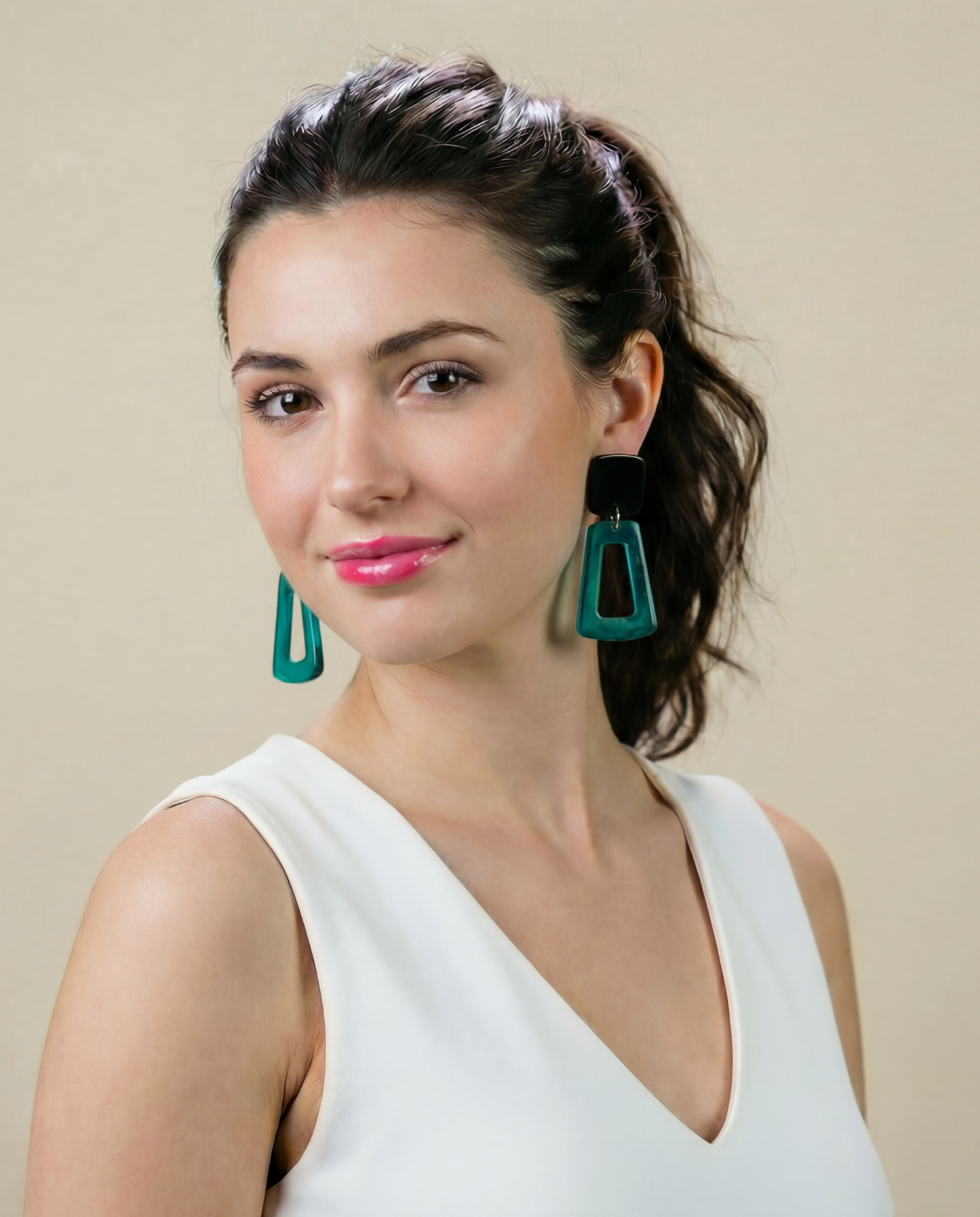 Woman wearing a white sleeveless top with teal earrings against a beige background